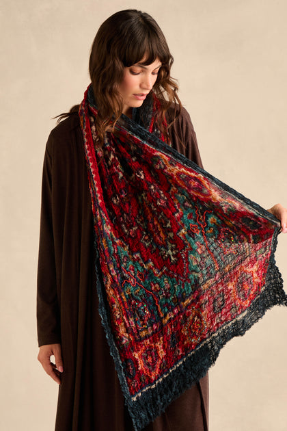 A person dressed in dark clothing holds the Crushed Sequin Detail Wool Scarf, showcasing its large size and intricate ruby, teal, and black design.