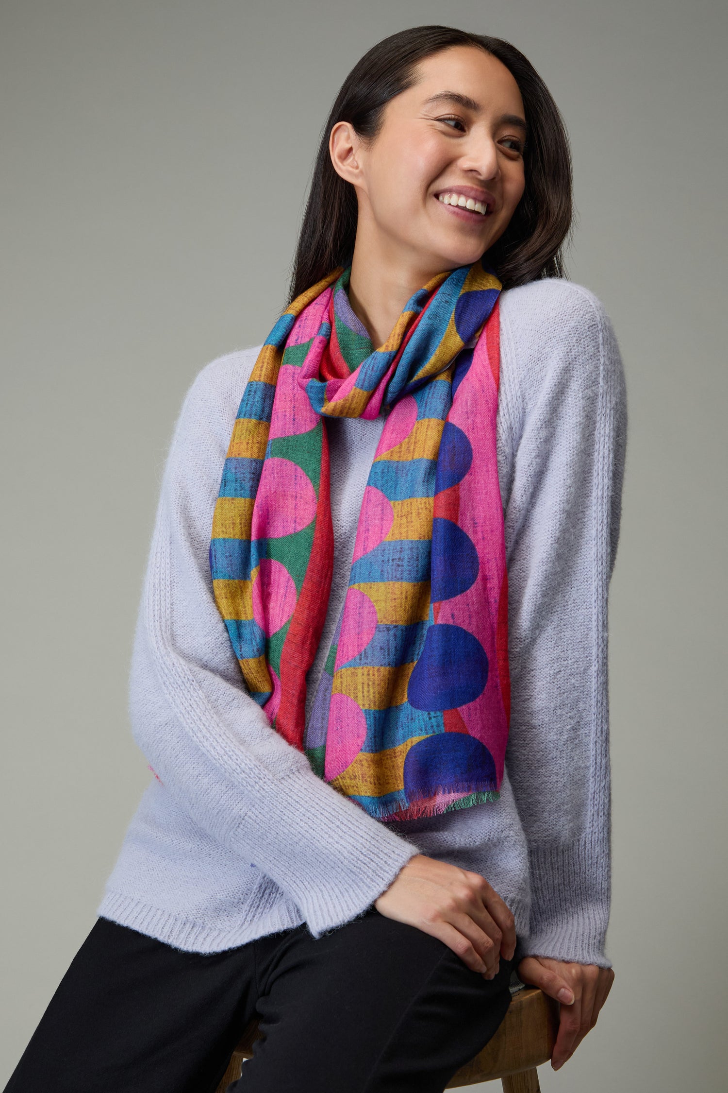 A woman sits on a stool, smiling and looking to the side, dressed in a light purple sweater, black pants, and the Colour Pop Circles Wool Scarf with vibrant polka dots.