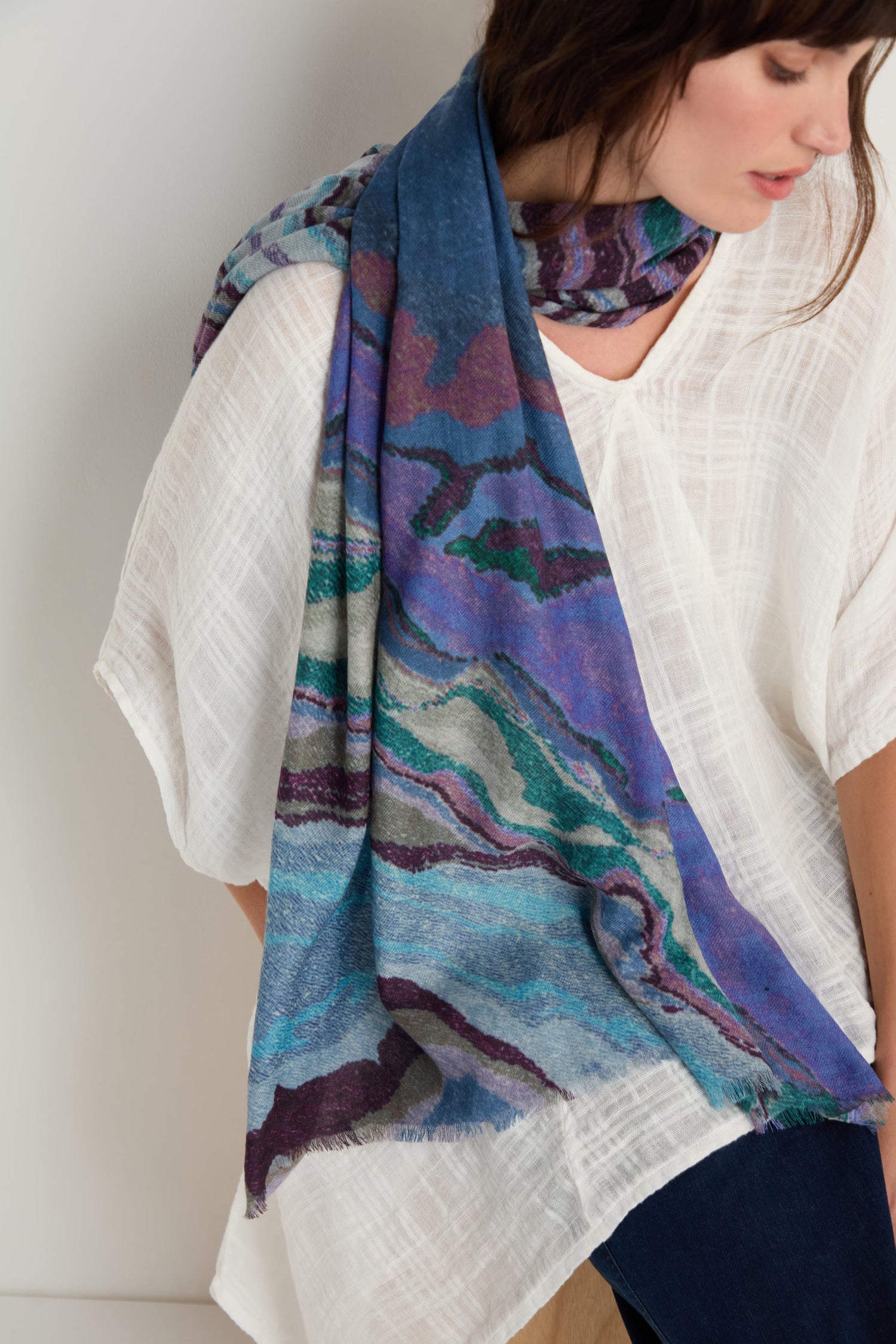 A woman stands against a plain light background, wearing a loose white textured top paired with the Agate Wool Scarf—a blue wool-silk accessory featuring a green and purple agate-inspired print.