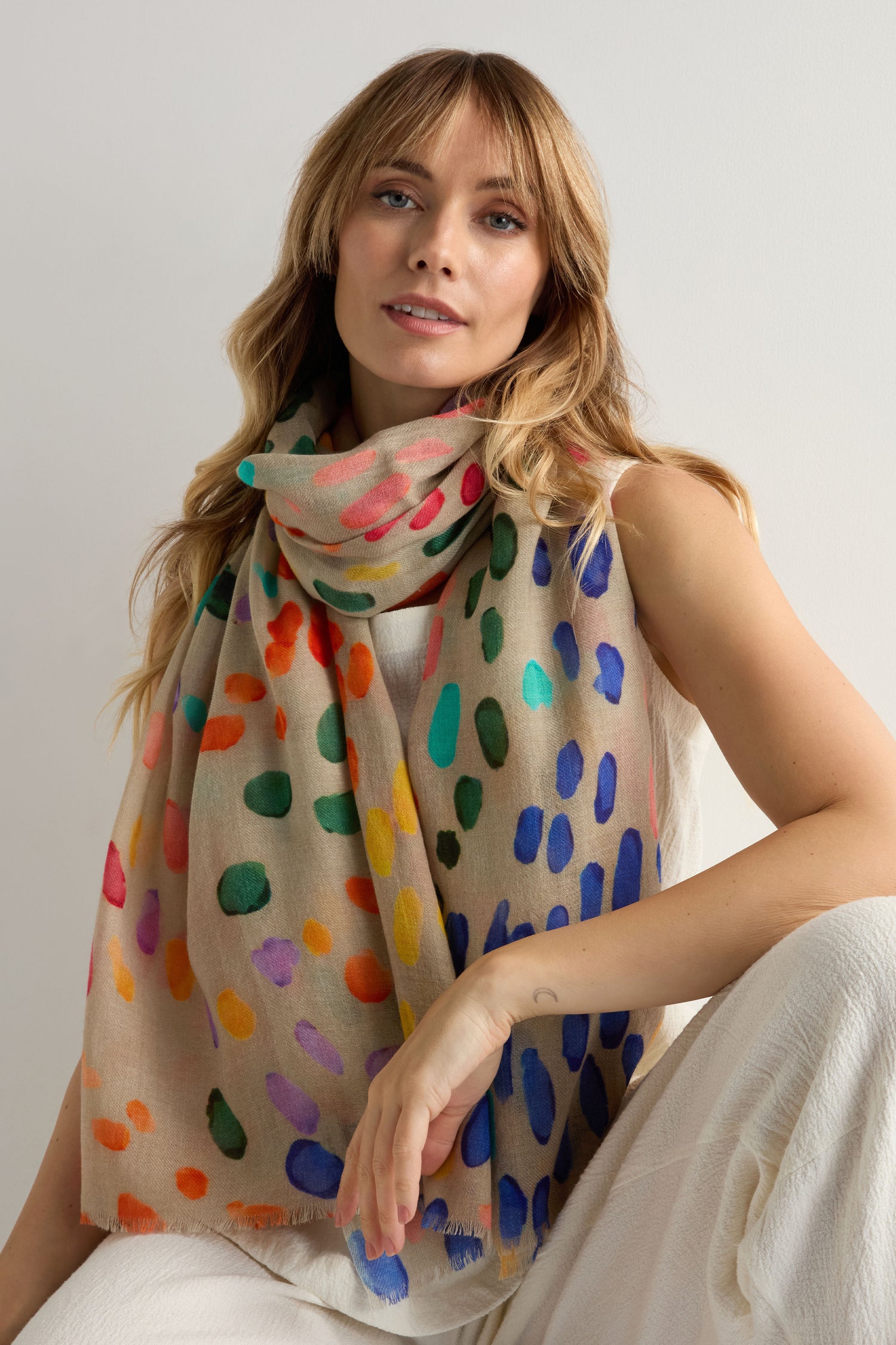 A person with long light brown hair wears a sleeveless white outfit and the Artful Dots Wool Silk Scarf, a lightweight accessory featuring colorful dot patterns, against a plain light background.