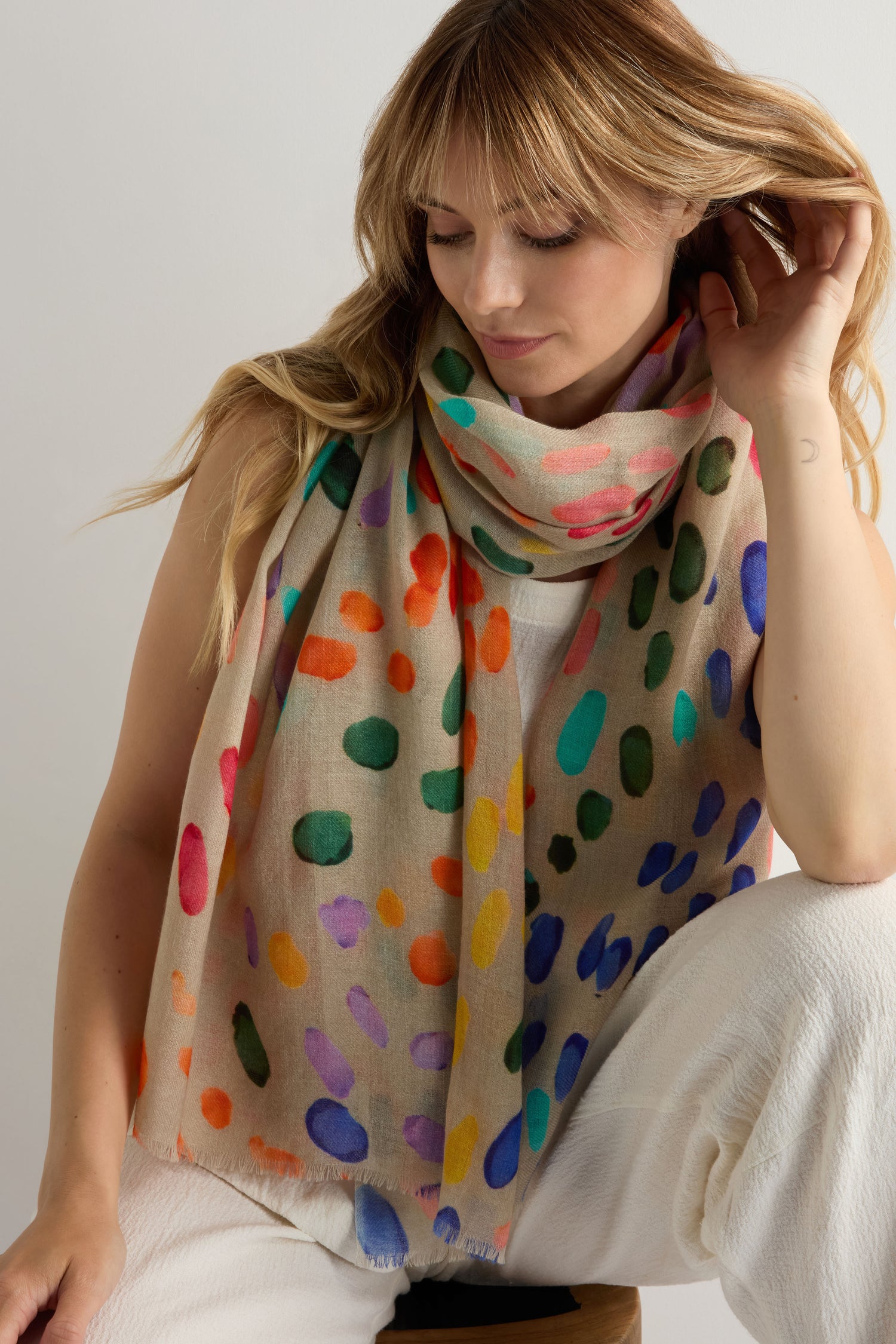 A woman sits on a stool, touching her hair, wearing the Artful Dots Wool Silk Scarf with multicolored spots over a white outfit.