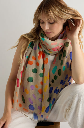 A woman sits on a stool, touching her hair, wearing the Artful Dots Wool Silk Scarf with multicolored spots over a white outfit.