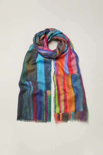 The Fluid Stripe Wool Scarf features a multicolored abstract pattern in blue, red, orange, green, and purple tones, shown against a plain background.