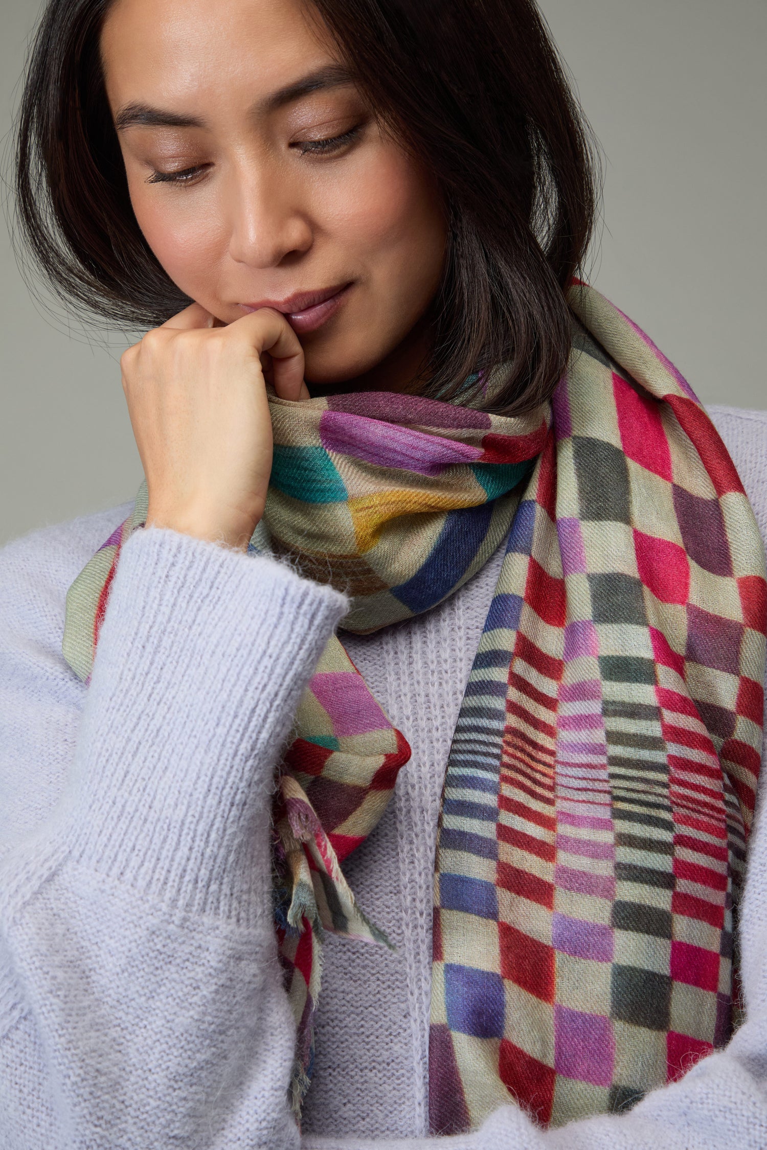 A person wearing a pastel purple sweater and the Mirage Check Wool Scarf looks down thoughtfully, resting their hand near their chin.