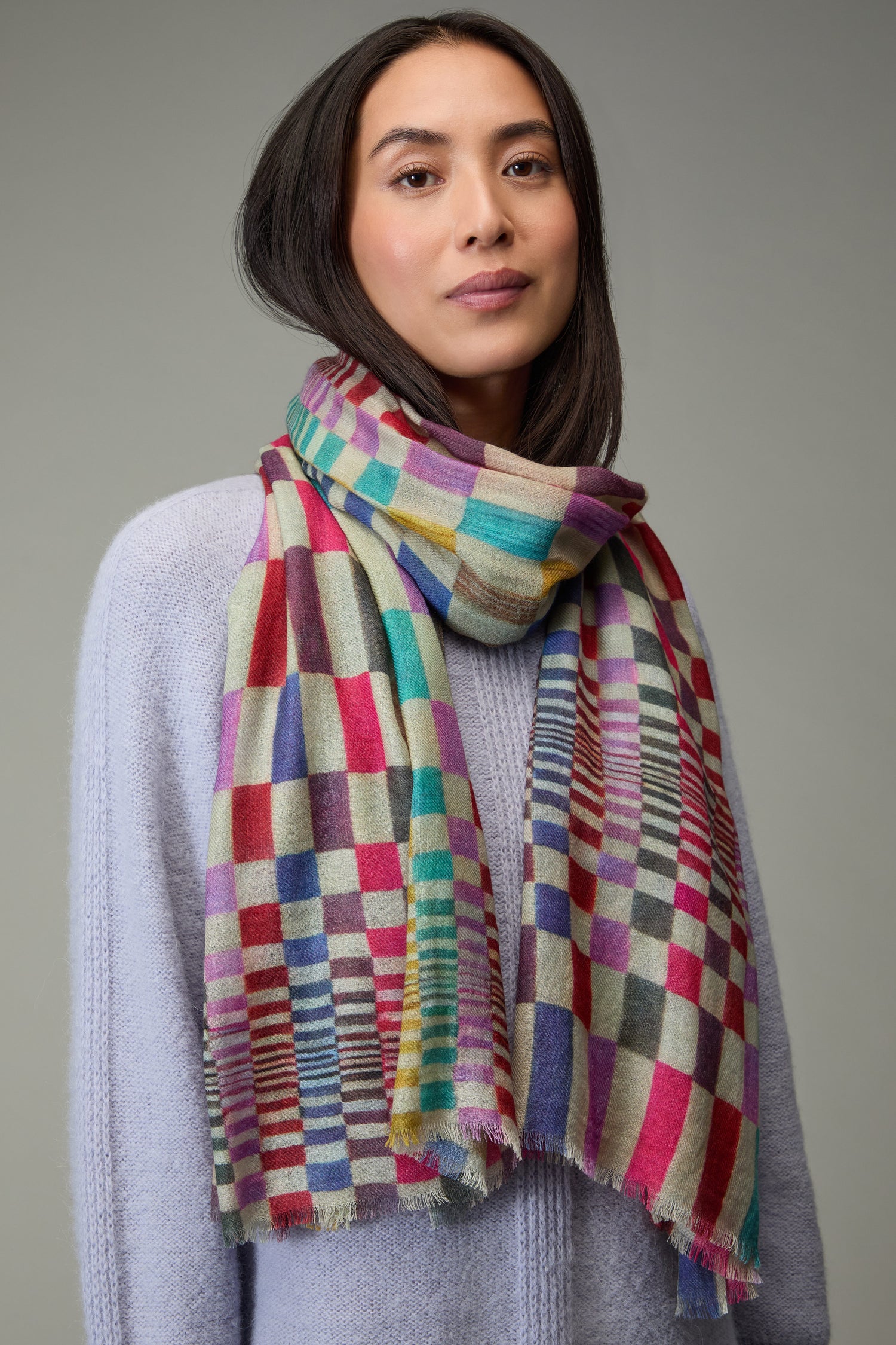 A woman models a Mirage Check Wool Scarf with a light purple sweater, standing against a plain grey background.