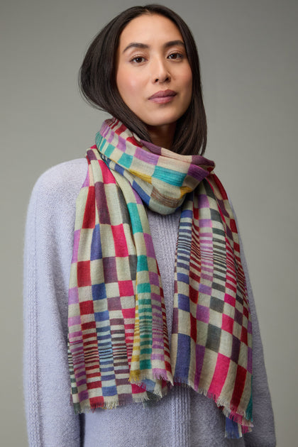 A woman models a Mirage Check Wool Scarf with a light purple sweater, standing against a plain grey background.