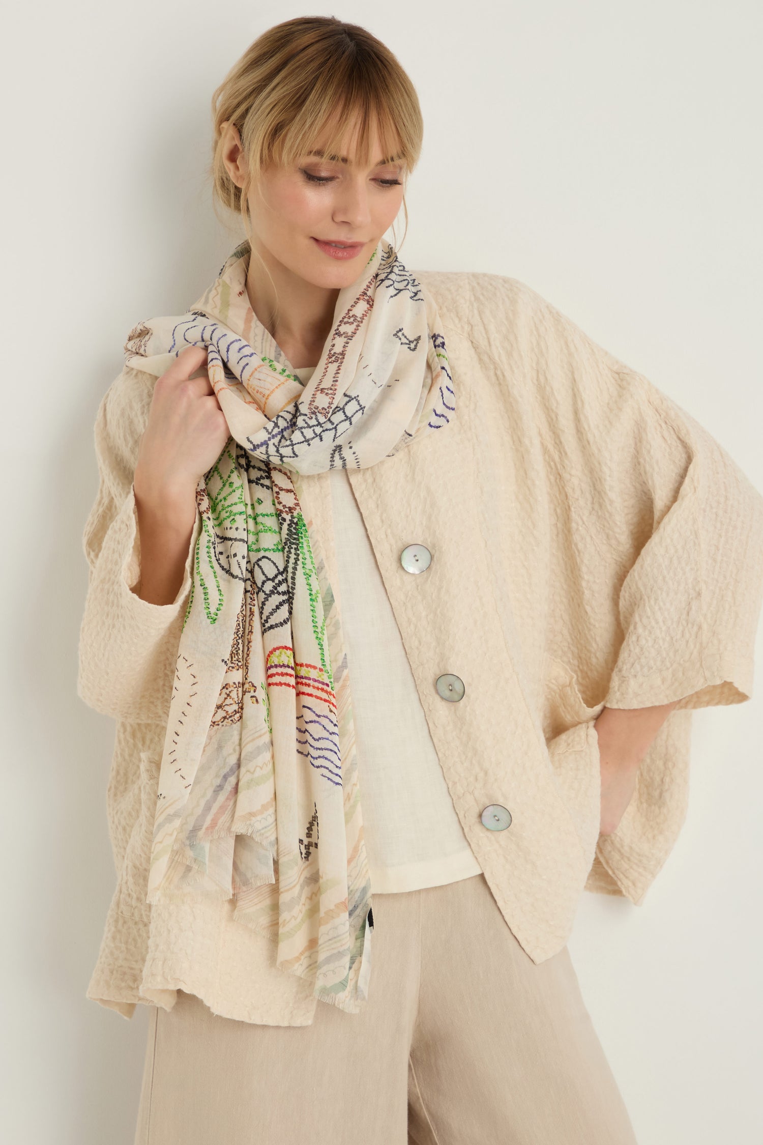 A woman stands against a plain off-white background wearing light pants, a textured beige jacket, and the By The Sea Cotton Modal Scarf featuring coastal motifs.