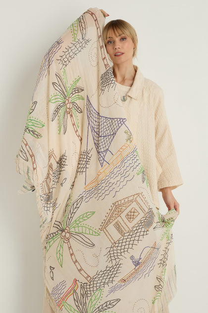 A woman in a light coat holds up the By The Sea Cotton Modal Scarf, a large, cream-colored accessory made from lightweight, breathable fabric with colorful coastal motifs such as palm trees, huts, and boats.