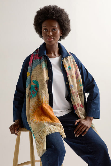 A person with an afro sits on a stool, dressed in a dark denim outfit and wearing the Riverside Cotton Mix Scarf, which adds floral and sun designs to their look. The lightweight cotton mix provides breathable comfort all day.