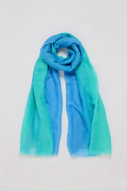 The Ombre Linen Mix Scarf, featuring shades of blue and turquoise in a gradient design, is a lightweight accessory that effortlessly enhances any outfit against its plain background.