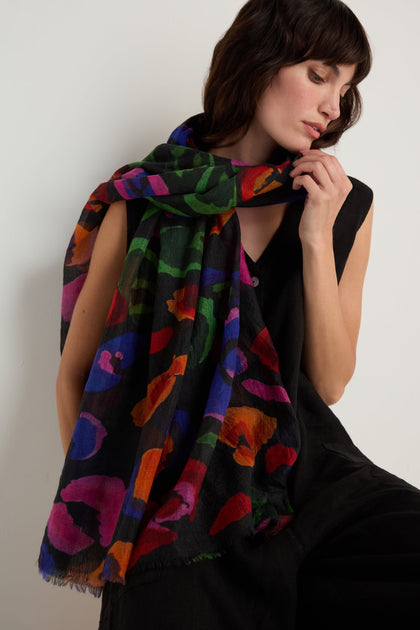 A woman wearing a sleeveless black outfit has the Painted Pansy Wool Scarf with a vibrant multicolour pansy print draped over her shoulders, looking down.