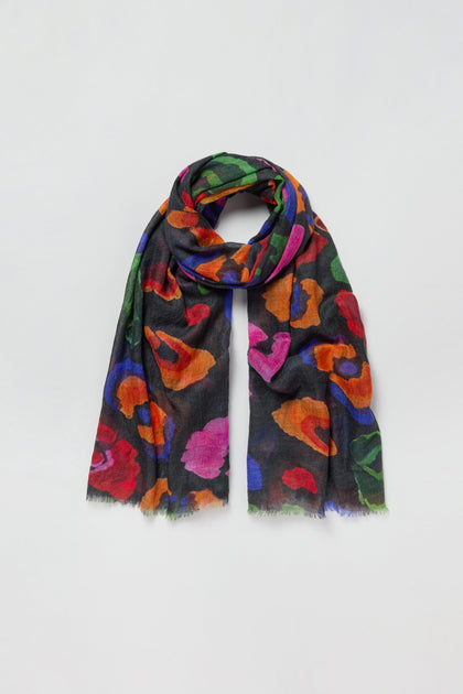 The Painted Pansy Wool Scarf is a black wool scarf adorned with vibrant floral patterns in red, orange, purple, and green, showcasing a bold painted pansy print. Displayed on a plain white background, this multicolour accessory is sure to brighten any outfit.