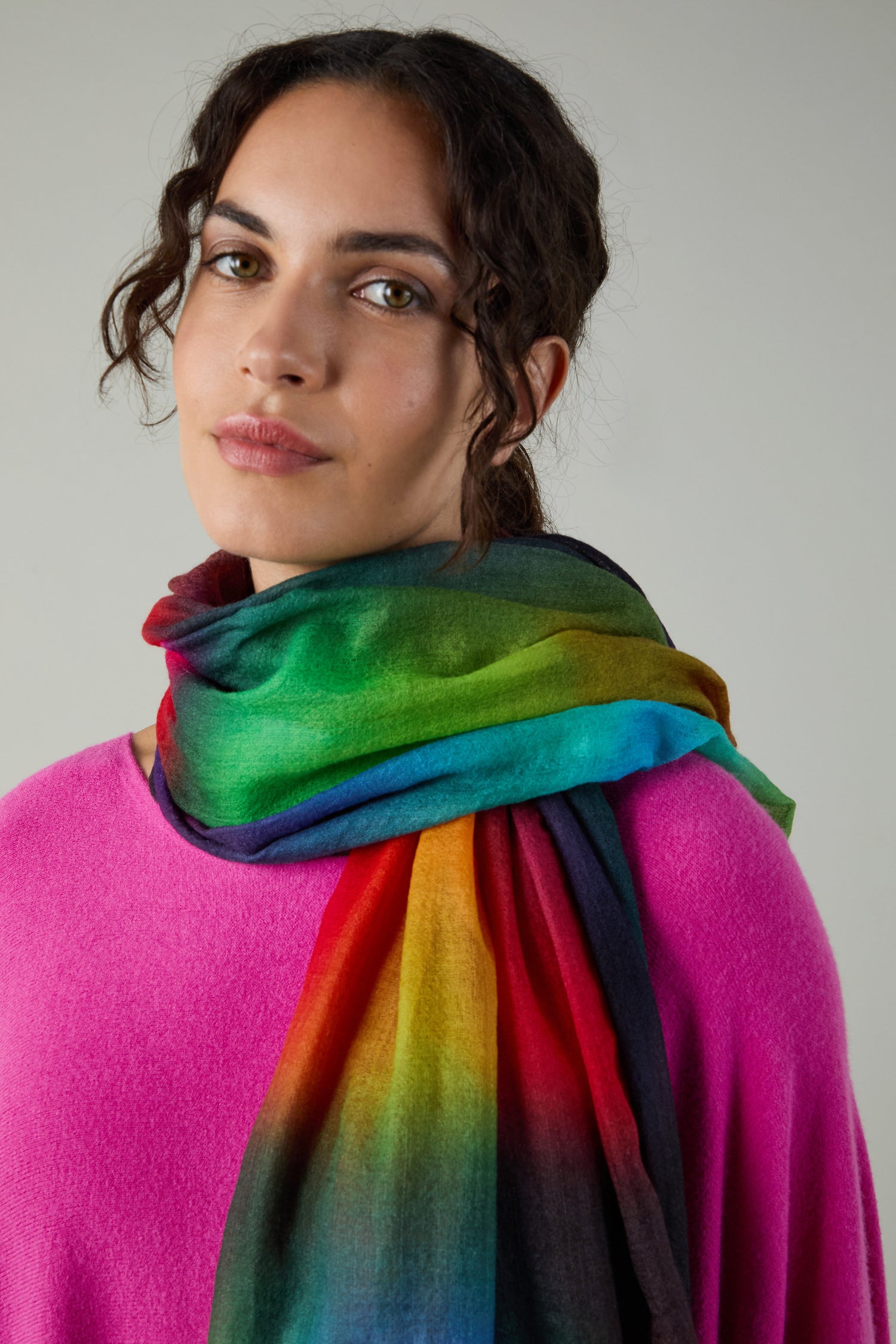 A person in a pink top and the Aurora Wool Scarf, featuring multicolored rainbow hues, looks at the camera against a plain background.