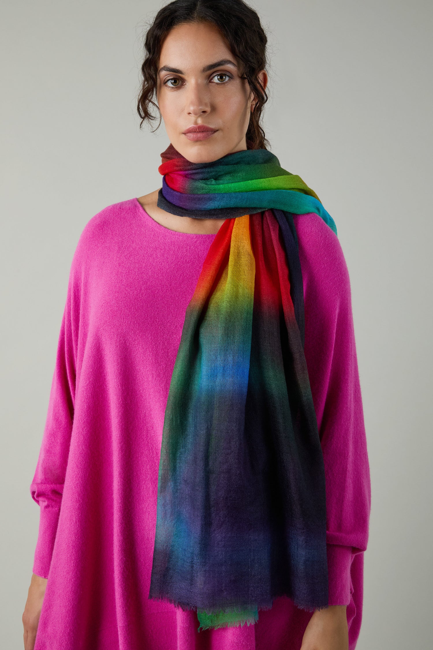 A woman in a vibrant pink sweater and the Aurora Wool Scarf, with its multicolored ombré design, stands against a simple light gray backdrop.
