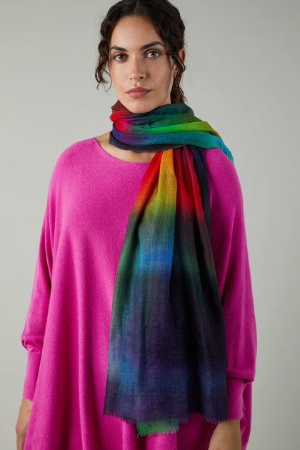 A woman in a vibrant pink sweater and the Aurora Wool Scarf, with its multicolored ombré design, stands against a simple light gray backdrop.