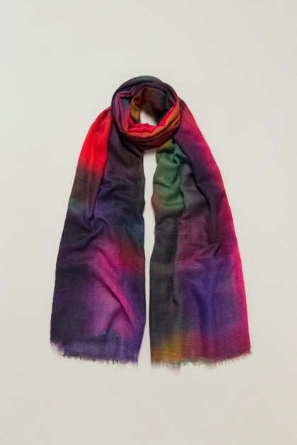 The Aurora Wool Scarf, a long and lightweight accessory featuring a red, green, blue, purple, and yellow gradient ombré pattern, is displayed on a plain background.