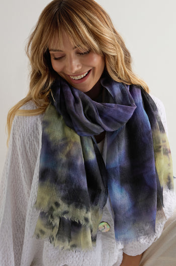 A woman smiles while looking down, wearing a white textured top and the Spiral Tie Dye Wool Scarf in vibrant blue, black, green, and yellow tones against a plain background.