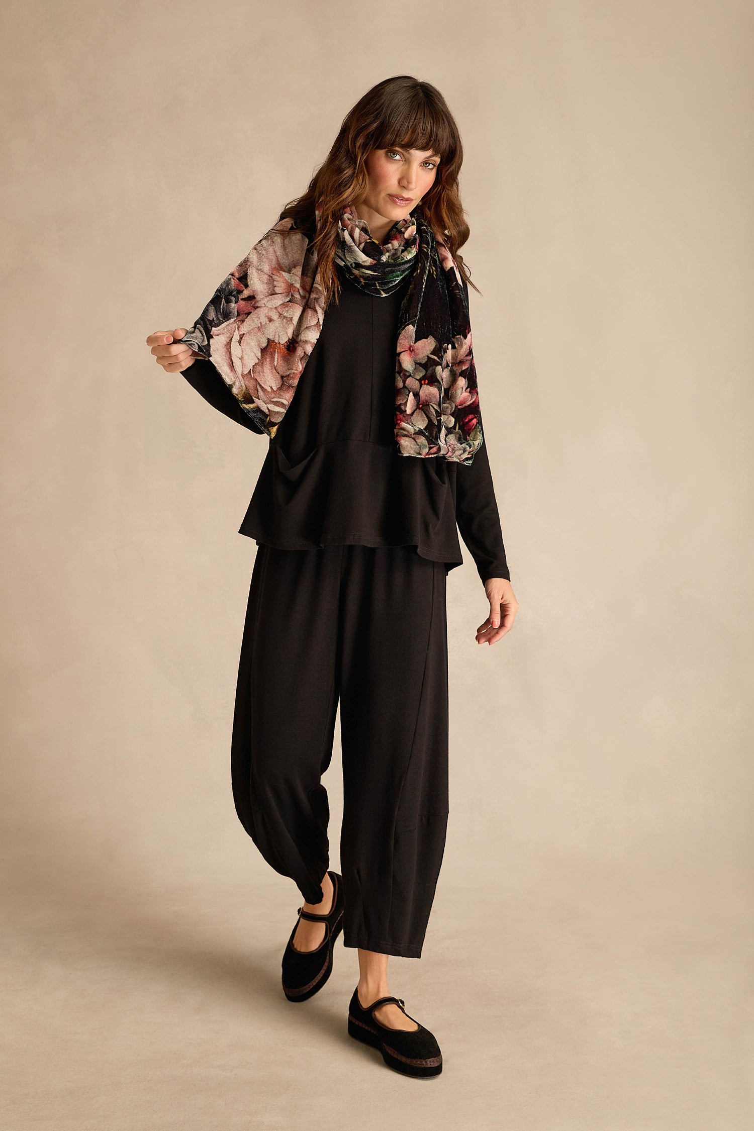 Woman standing against a neutral background, wearing a black outfit with loose pants, long sleeves, black shoes, and the Winter Rose Velvet Scarf featuring a delicate winter rose print draped around her shoulders.