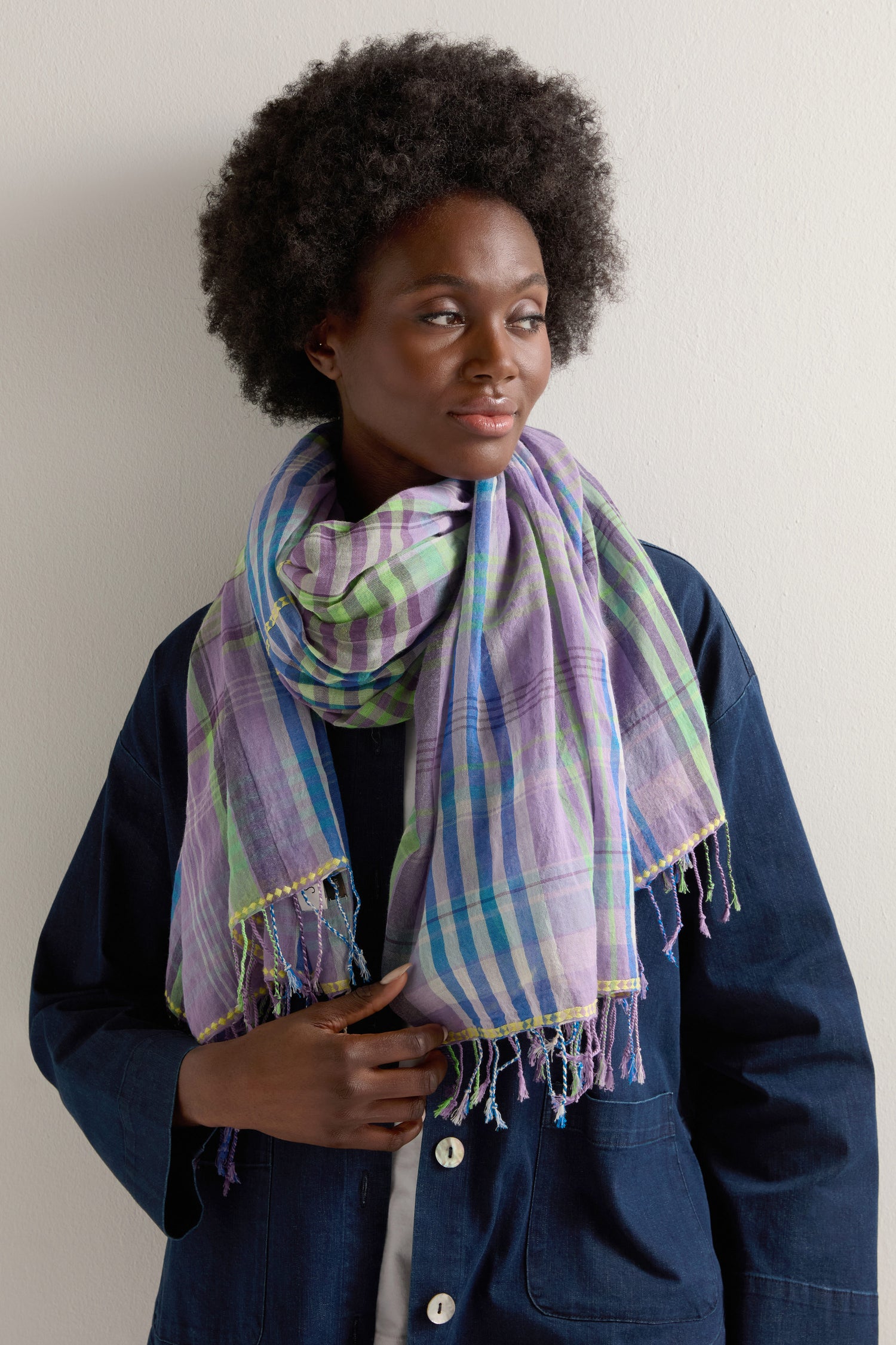 A person wears a Check Stitch Cotton Scarf, featuring vibrant purple, green, and blue stripes, over a dark blue jacket against a light-colored wall. This accessory adds a burst of color to their look.