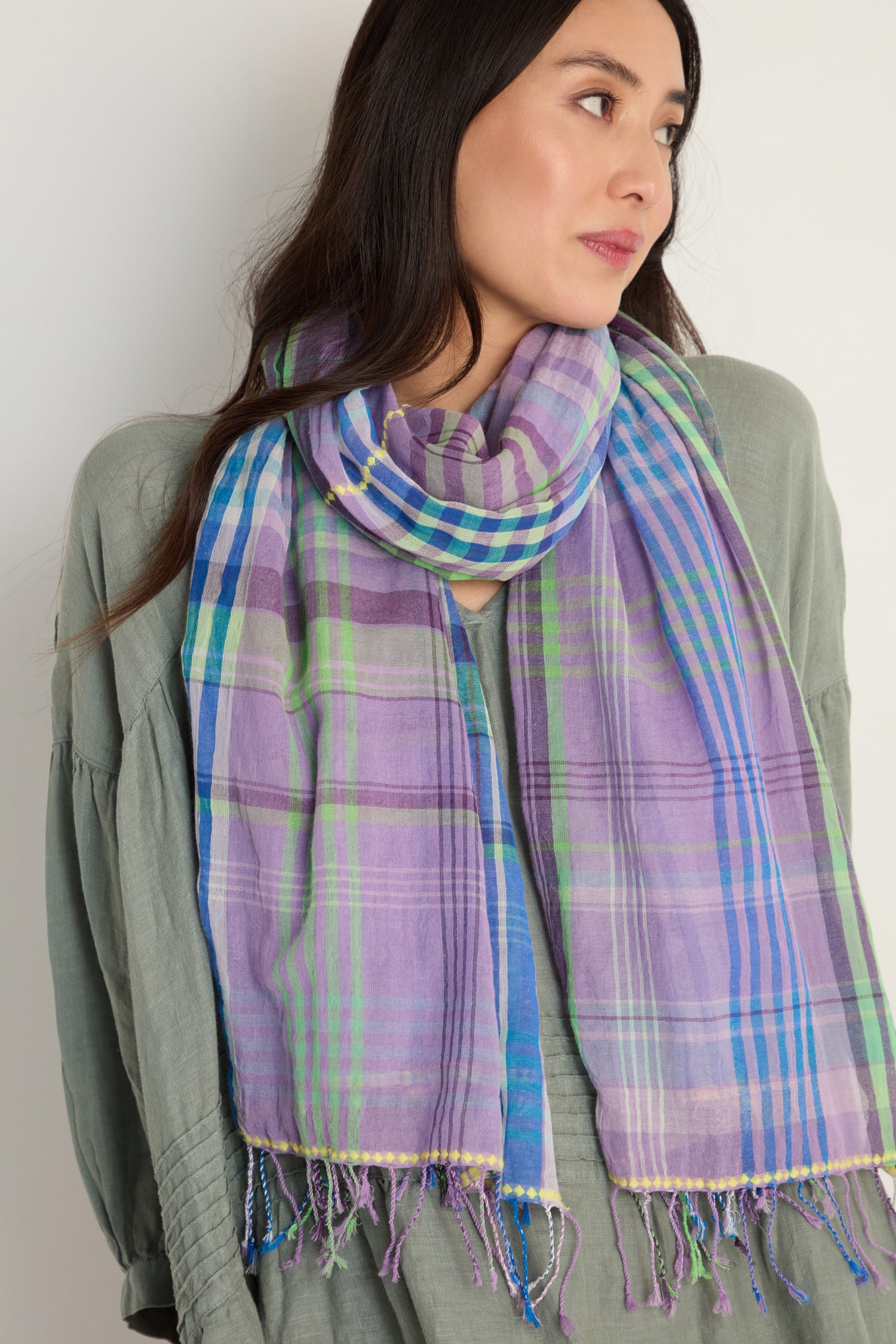 A woman wears a light green top and the Check Stitch Cotton Scarf, a lightweight cotton accessory in purple, blue, and green plaid with fringed ends, looking slightly to the side against a plain background.