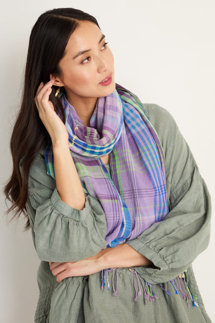 A woman wearing a loose green top and the Check Stitch Cotton Scarf with a purple plaid pattern stands against a plain background, looking to the side and touching her hair.