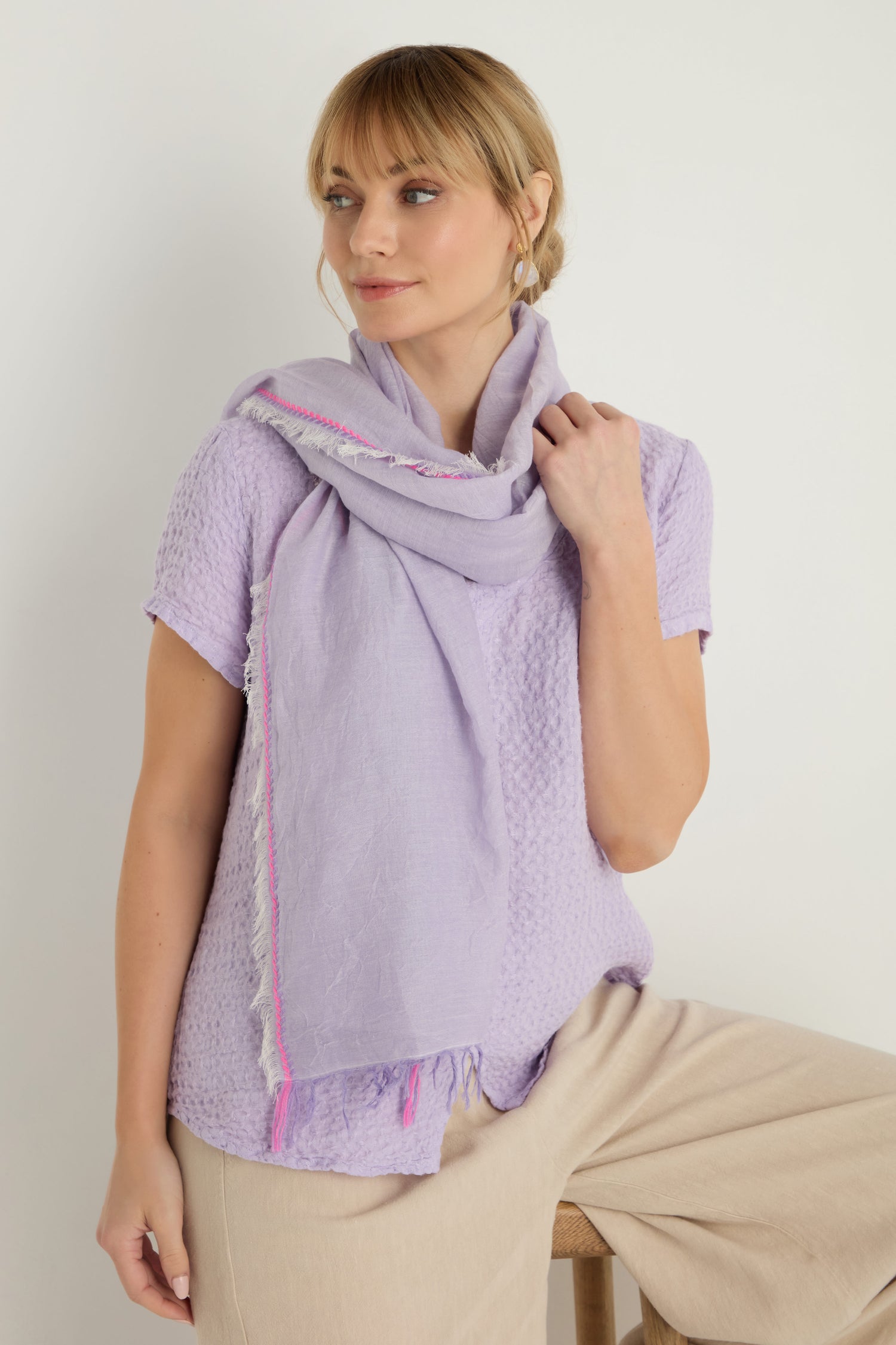 A woman sits on a stool against a plain background, wearing beige pants, a light purple textured top, and the Lilac Fringe Cotton Scarf—a cotton scarf in lilac with pink accents.