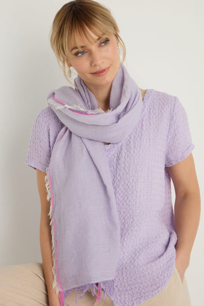 A woman stands against a plain light background wearing a textured light purple short-sleeve top and the Lilac Fringe Cotton Scarf.