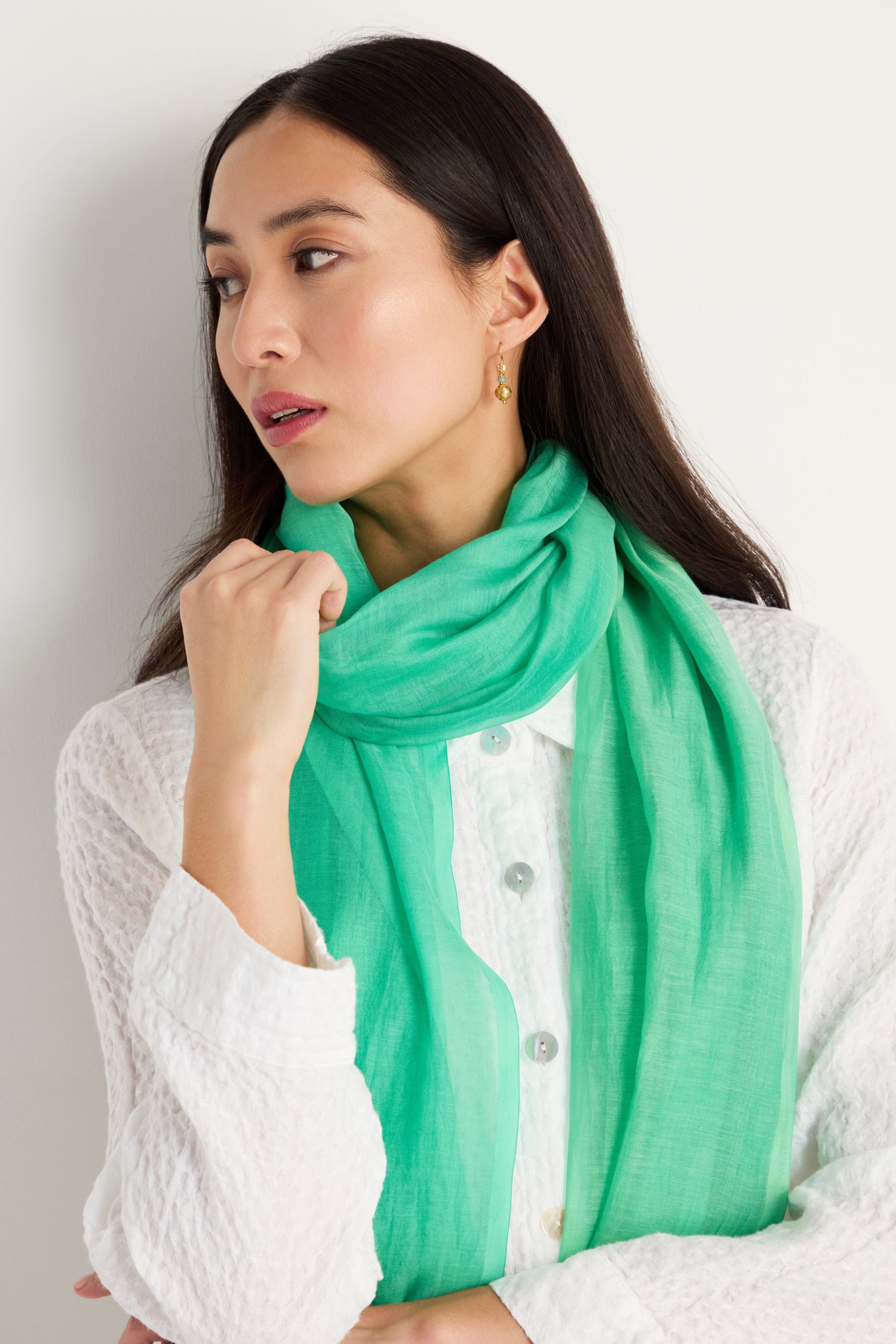 A woman wears a textured white button-up top and the Verdant Cotton Blend Scarf in bright green, looking to the side with her hand near her neck against a plain background.