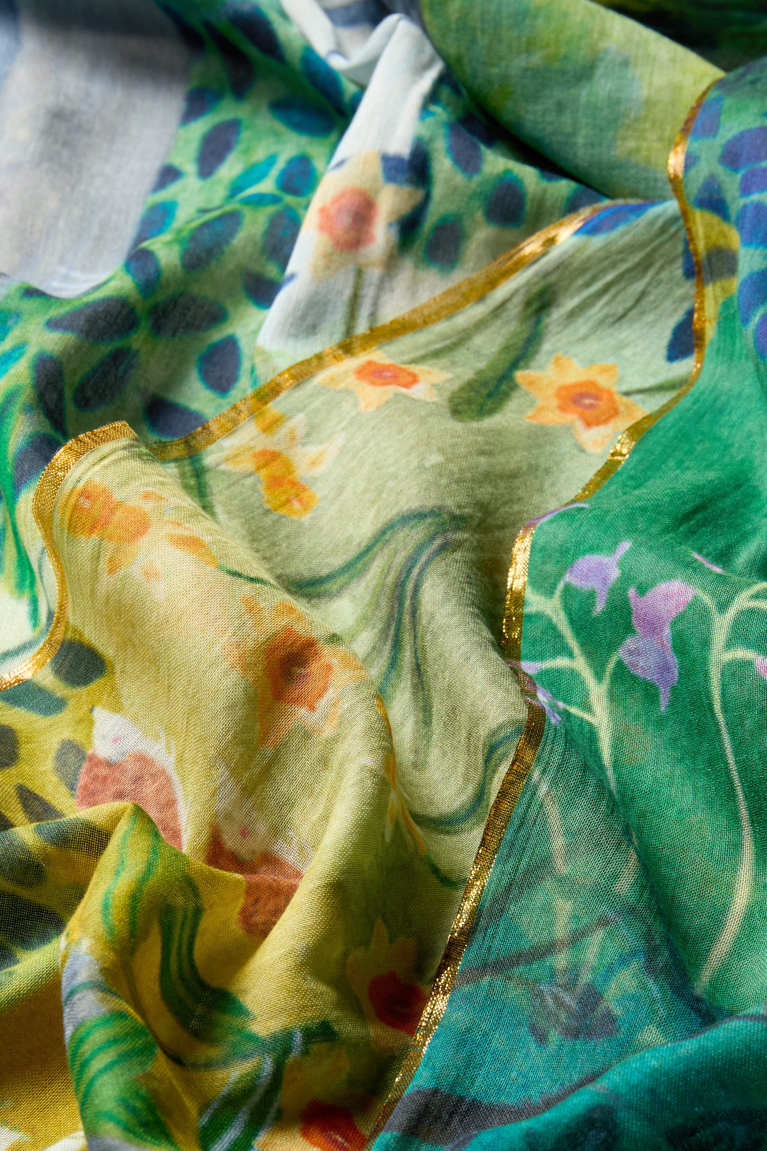 A close-up of the Painted Garden Cotton Scarf reveals vibrant floral patterns in shades of green, yellow, and blue with gold trim. This lightweight and versatile accessory exudes artistic elegance.