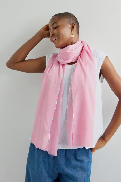 A person in a sleeveless white top, blue pants, and the versatile Silver Edge Cotton Scarf smiles while posing against a plain white wall.