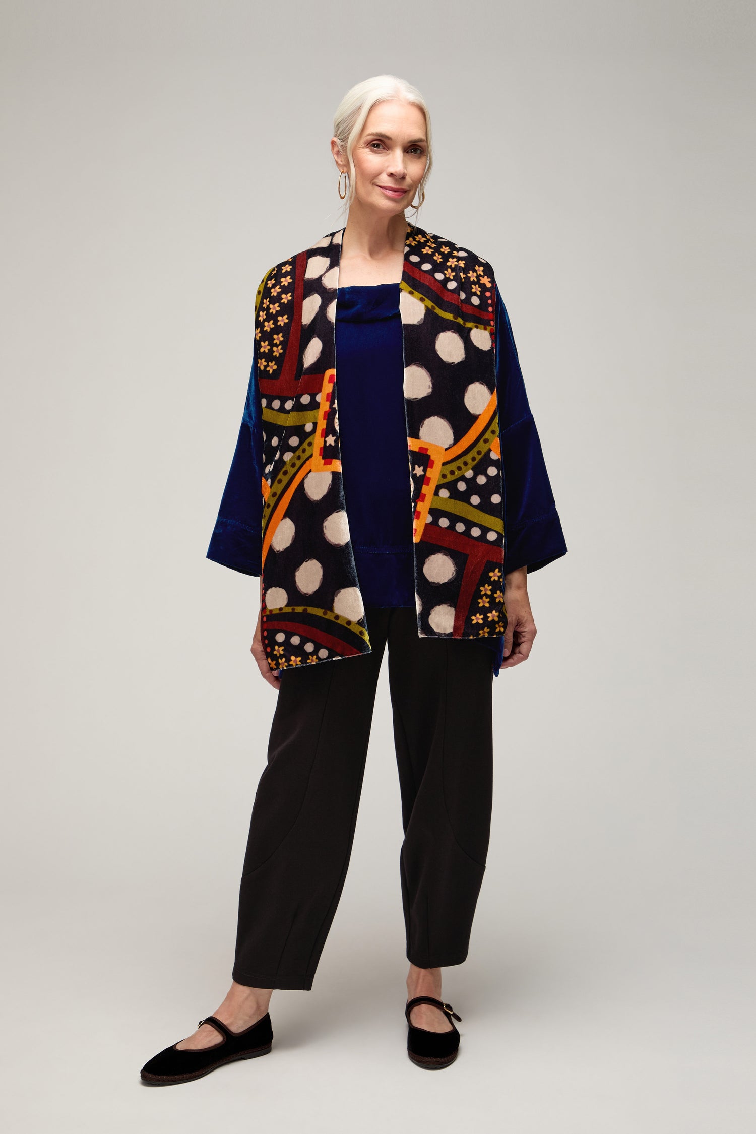Older woman with white hair stands in a studio wearing a blue top, black pants, black shoes, and the Hera Velvet Scarf with colorful traditional motifs and polka dots.