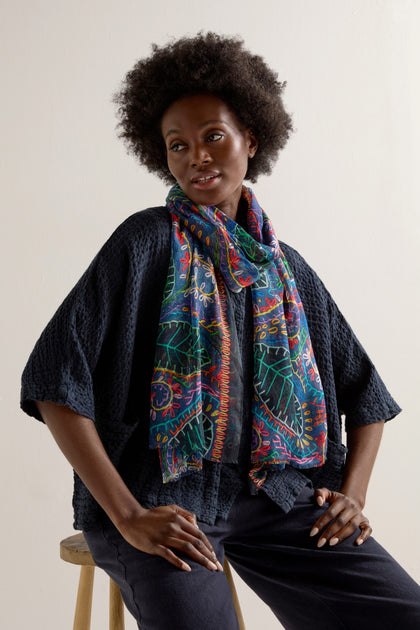 A person with an afro hairstyle sits on a stool in a dark textured outfit, accessorized with the Painted Leaves Modal Scarf, showcasing a luxuriously soft modal fabric adorned with a hand-painted leaf design.