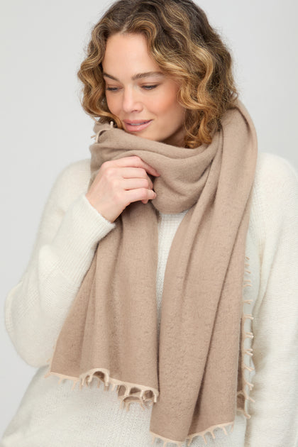 A person with curly hair wears a white sweater and the Contrast Trim Cream Cashmere Scarf, looking downward with a slight smile.