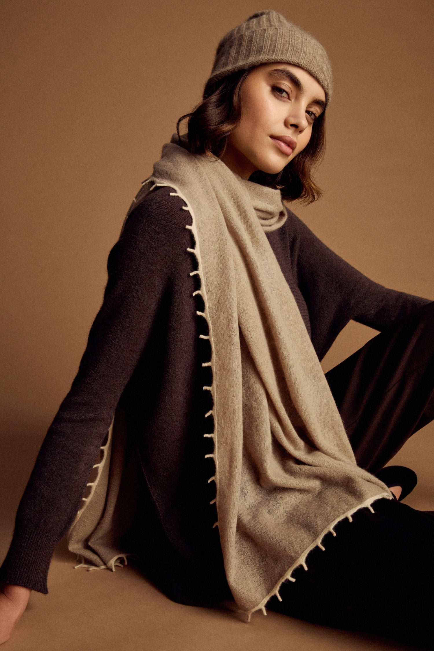 A woman wearing a Contrast Trim Cream Cashmere Scarf and knit hat with a black long-sleeve top sits against a tan background, looking at the camera with a relaxed expression.