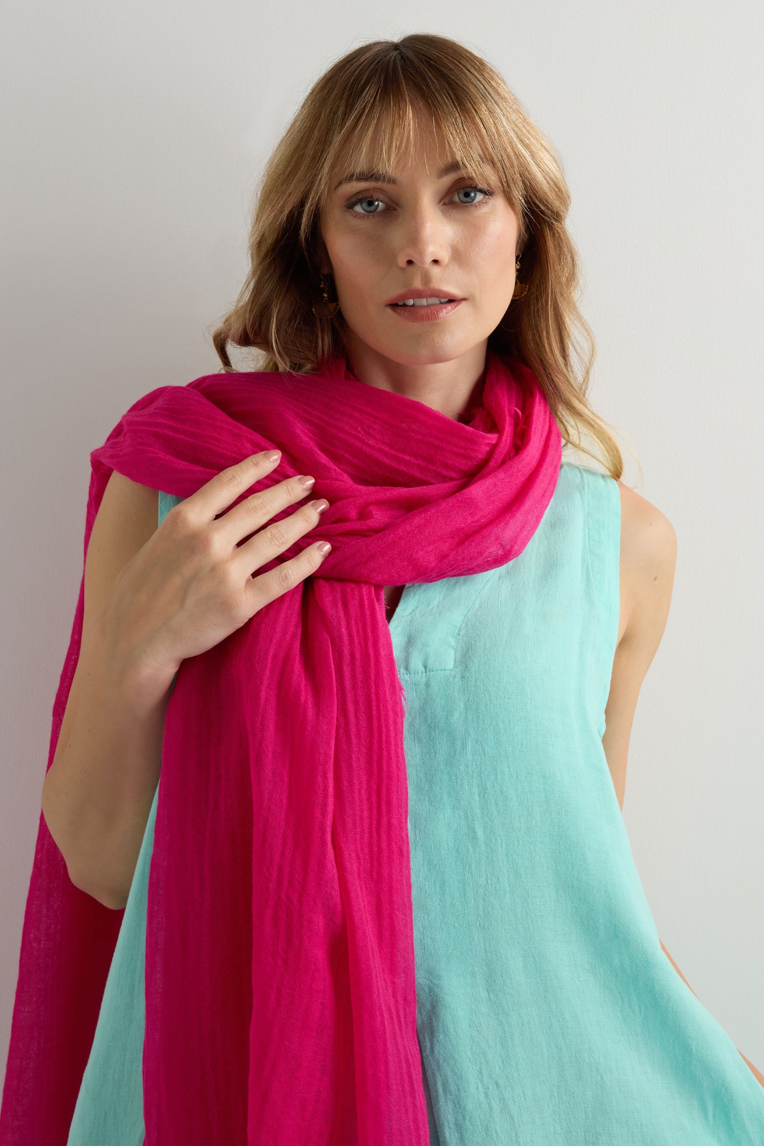 A woman wearing a light blue sleeveless top and the Hibiscus Fine Wool Fringe Scarf draped over her shoulders stands against a plain light-colored wall.