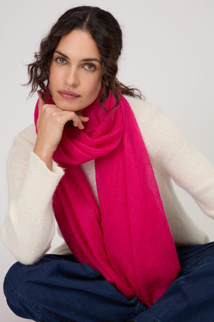 Woman with wavy dark hair in a white sweater and blue jeans, wearing the Hibiscus Fine Wool Fringe Scarf in fuchsia, sits with one hand resting against her face.