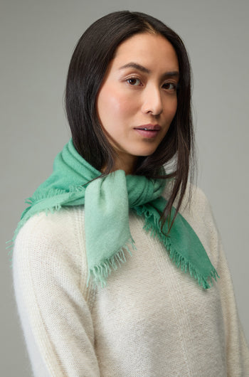 Woman with long dark hair wears a white sweater and the Ombre Cashmere Square Scarf in light green, standing against a plain grey background.