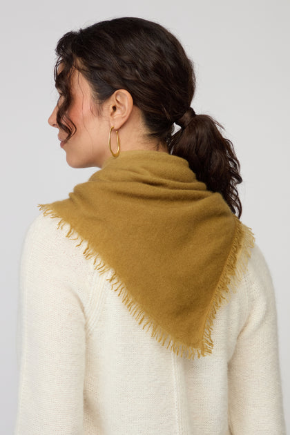 A woman with dark hair in a ponytail, wearing gold hoop earrings, a white sweater, and the Ombre Cashmere Square Scarf in mustard yellow draped over her shoulders, faces away from the camera.