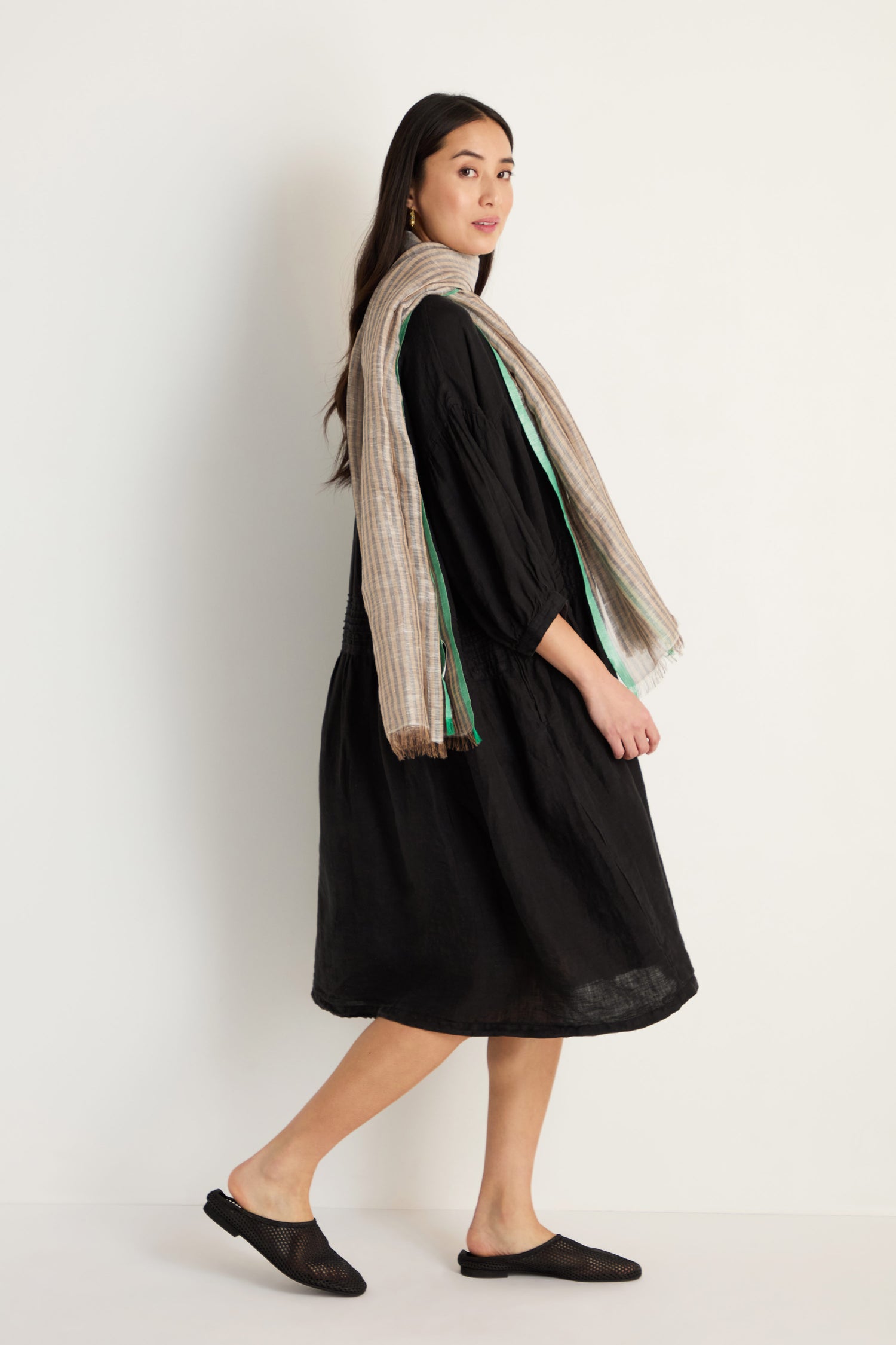 A woman stands sideways against a white background, wearing a black dress, black slip-on shoes, and the Spearmint Trim Soft Stripe Scarf that adds a fresh touch to her outfit.