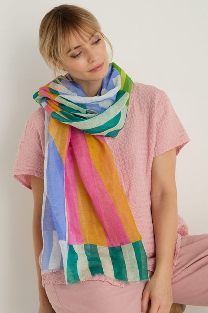 A person stands against a plain light background, wearing a light pink textured shirt and pants, accessorized with the Sunburst Block Print Linen Blend Scarf draped around their neck.