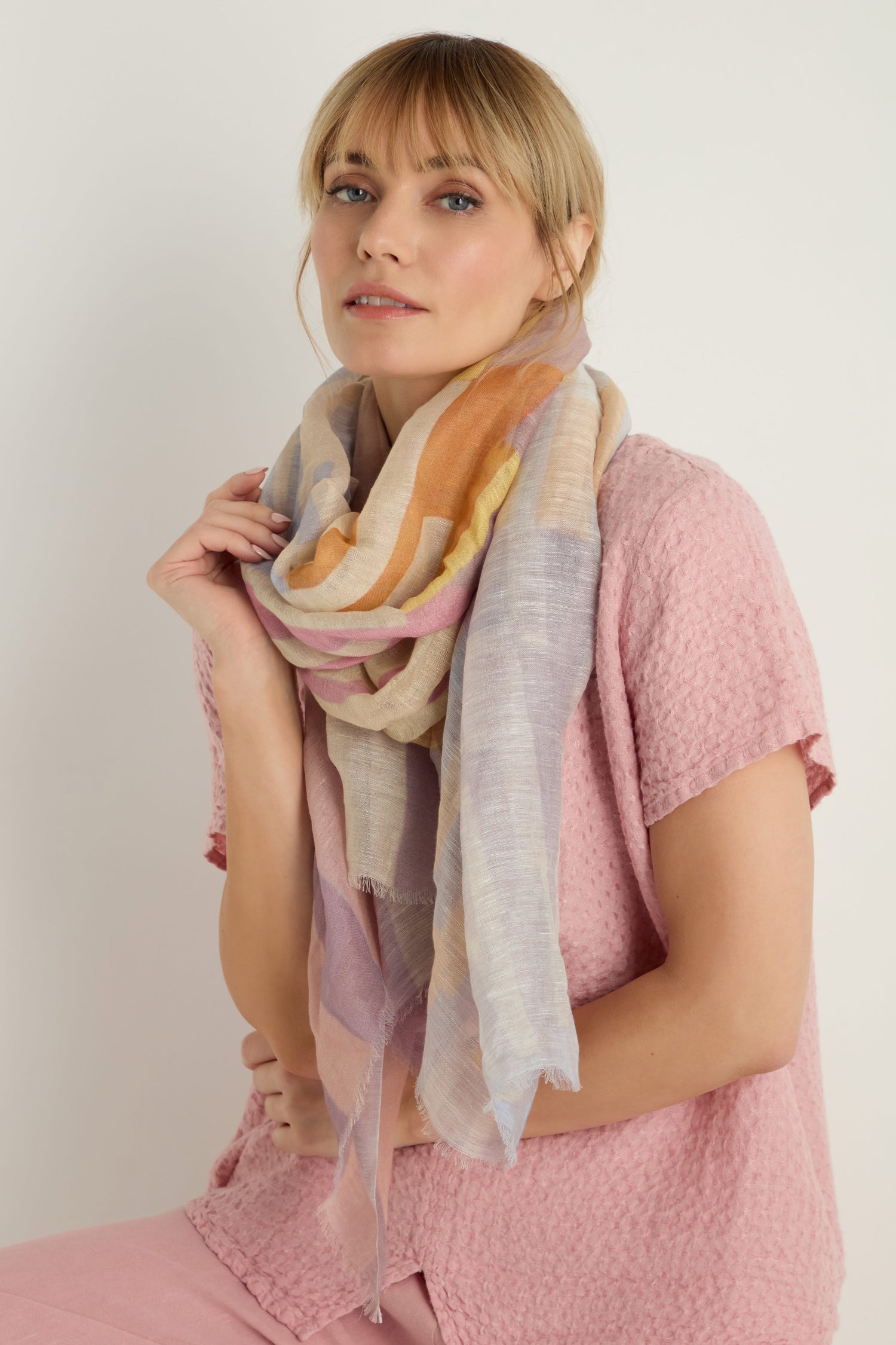 A woman in a textured pink top faces the camera, wearing the Soft Stripe Linen Blend Scarf in pastel colors around her neck and holding one end with her hand.