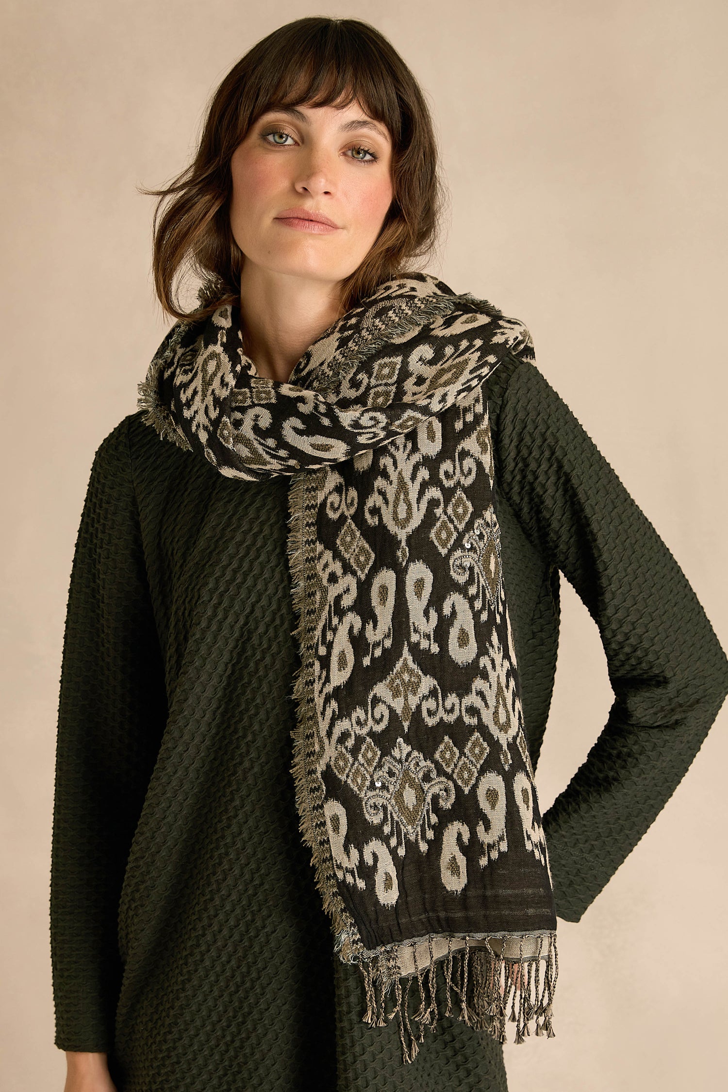 A woman stands against a neutral background wearing a textured dark green dress made from a cozy cotton-wool blend, styled with the Motif Sequin Scarf—a patterned black and white scarf featuring fringed ends.