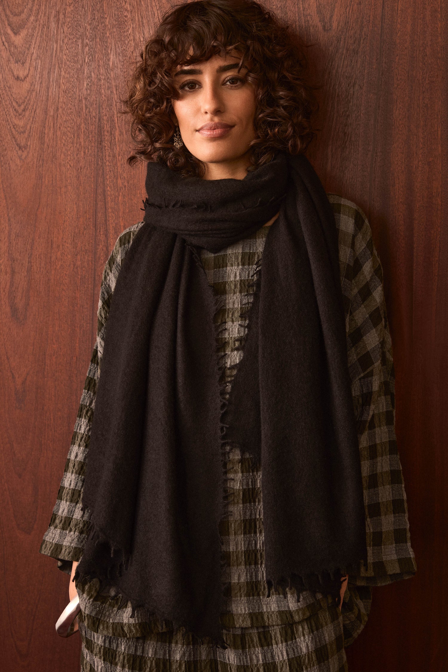 A woman with curly hair stands against a wooden wall, wearing a checkered dress and casually holding a smartphone in her left hand, styled with the Black Soft Cashmere Scarf draped over her shoulders.