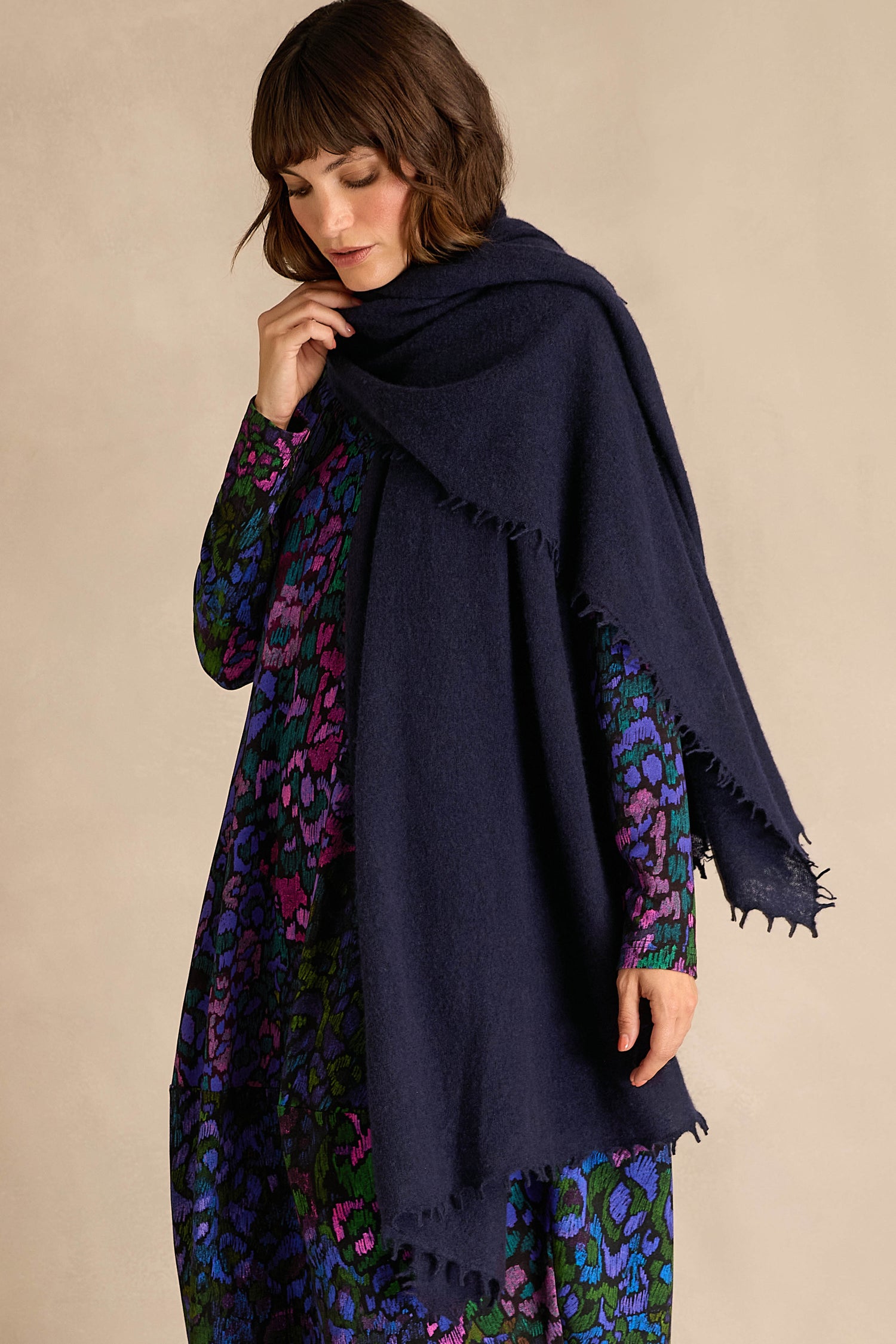 A person with short brown hair wears the Navy Soft Cashmere Scarf draped over a long-sleeve, patterned dress featuring purple, blue, and green tones, standing against a beige background.