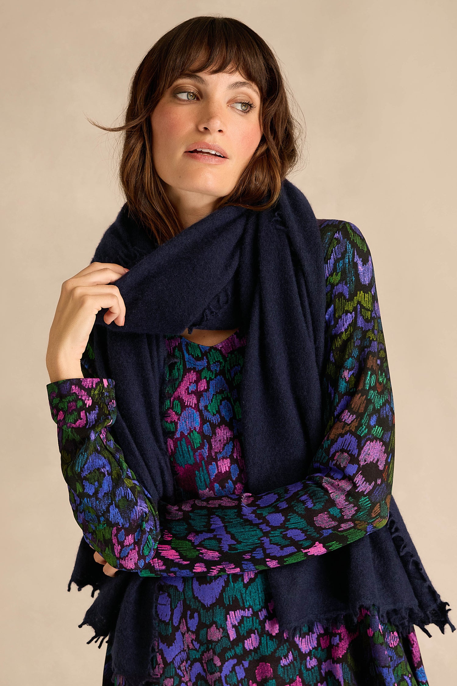 A woman wearing a patterned dress in shades of blue, green, and purple accessorizes her look with the Navy Soft Cashmere Scarf, standing against a plain beige background.