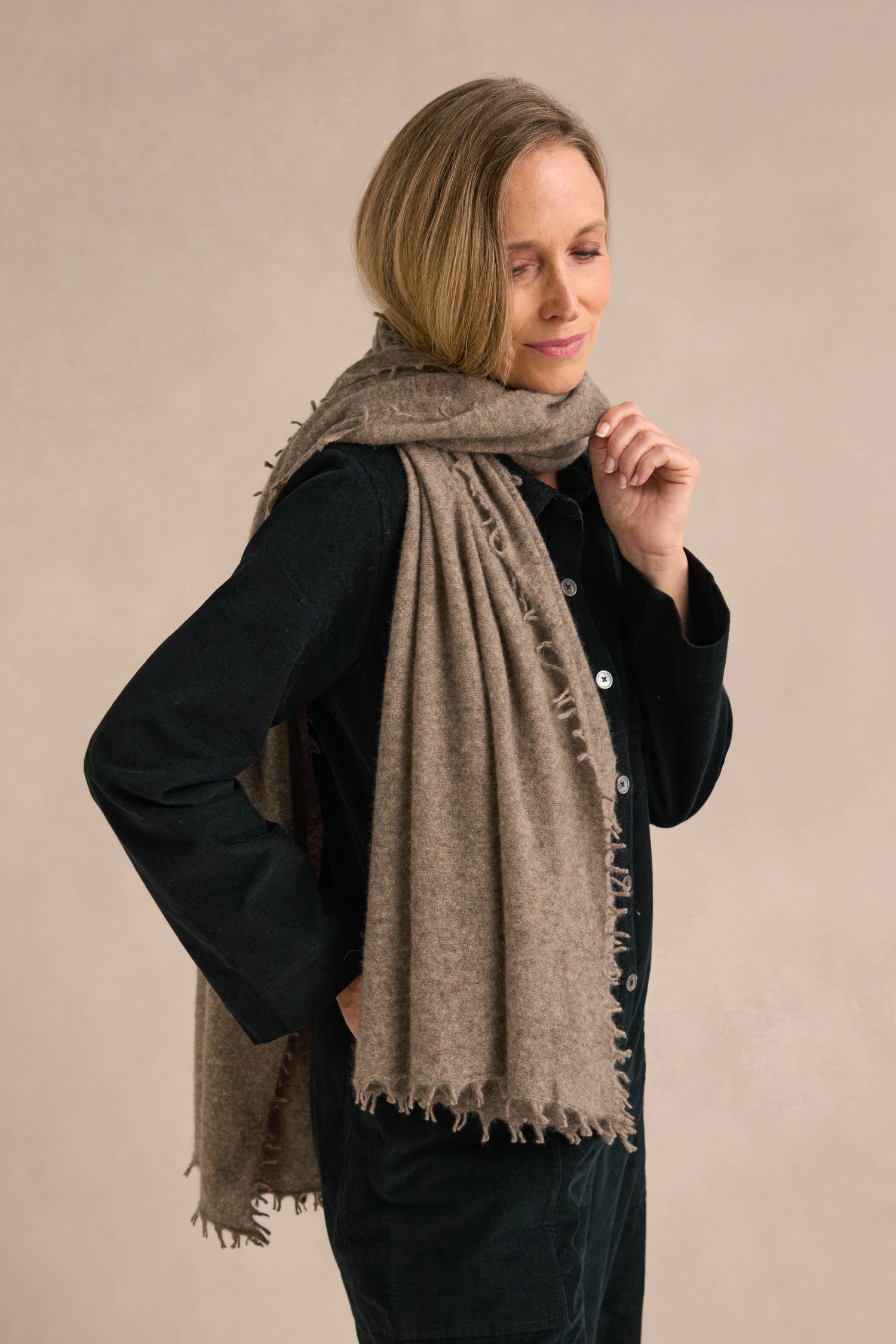 A person with short blonde hair wears a black outfit and the Sand Soft Cashmere Scarf, a large, textured accessory crafted from soft cashmere, while standing against a plain light beige background.