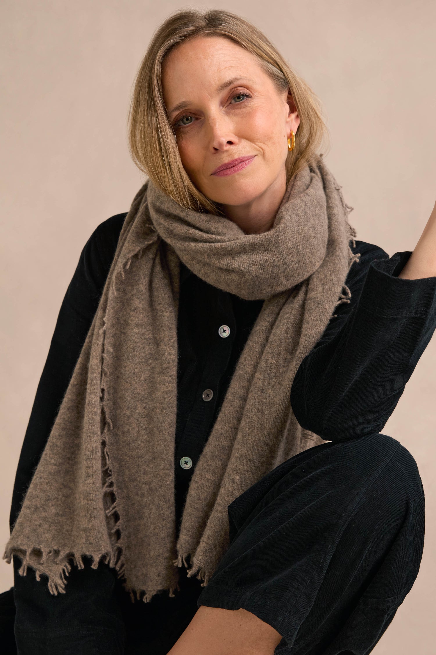 A woman with shoulder-length blonde hair wears a black outfit and the Sand Soft Cashmere Scarf, lounging with one arm resting on her knee against a neutral background.