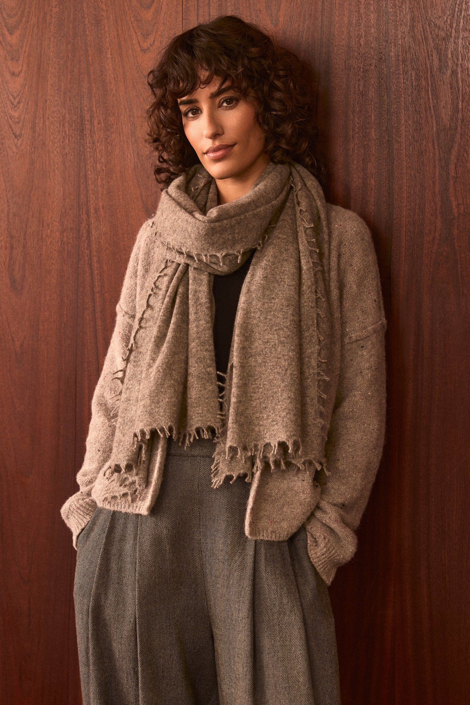 A person with curly brown hair stands against a wooden wall, wearing the Sand Soft Cashmere Scarf, a beige cardigan, and gray high-waisted pants, with hands in their pockets and a neutral expression.