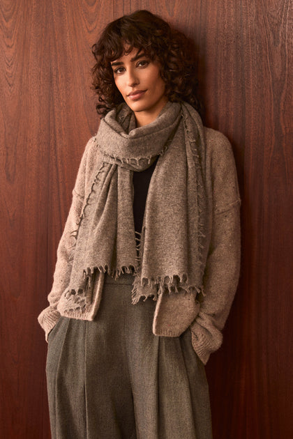 A person with curly brown hair stands against a wooden wall, wearing the Sand Soft Cashmere Scarf, a beige cardigan, and gray high-waisted pants, with hands in their pockets and a neutral expression.