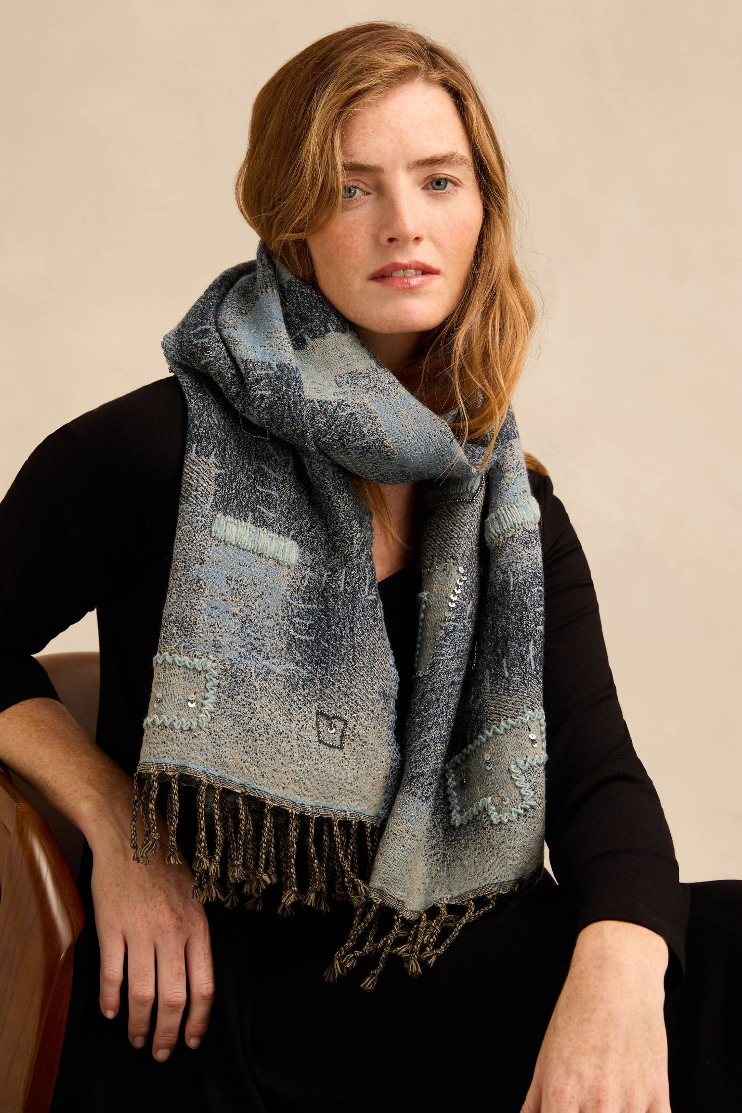 A woman with long, light brown hair is seated on a chair, looking at the camera with a neutral expression, wearing a large Etched Patchwork Sequin Wool Scarf in blue over a black outfit.